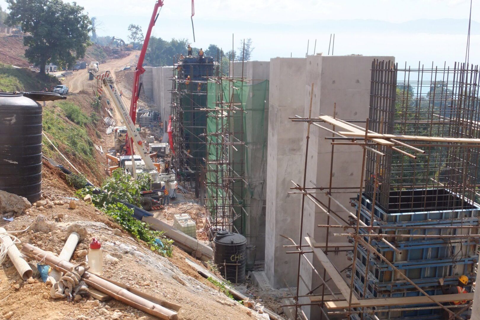Construction site with concrete foundation walls, rebar, scaffolding, and a red crane beside a dirt embankment
