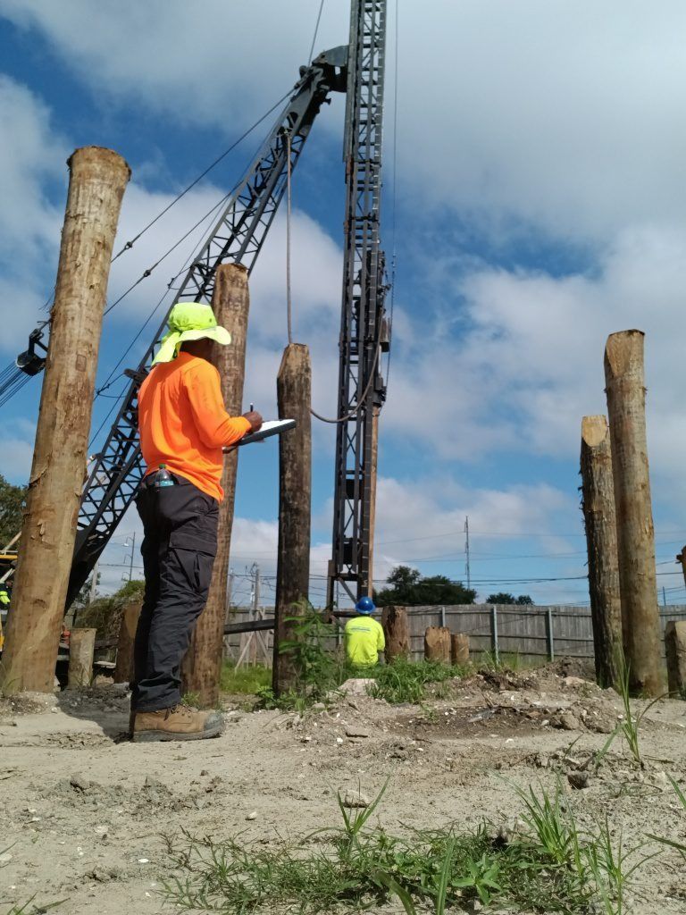 Construction workers in orange vests and hard hats at a drilling site with piles and heavy equipment