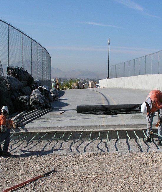 Workers laying concrete on a curved path beside fenced sports courts on a sunny day