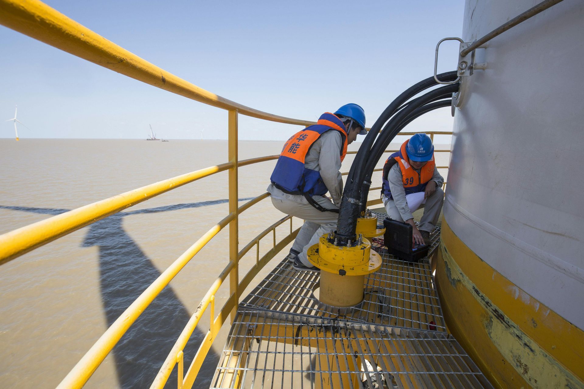Two workers in orange safety vests and blue helmets inspect equipment on a yellow industrial platform.