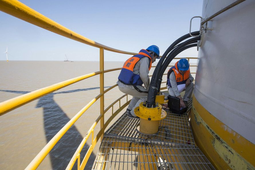 Two workers in orange vests on a yellow industrial platform beside large pipes, overlooking the water