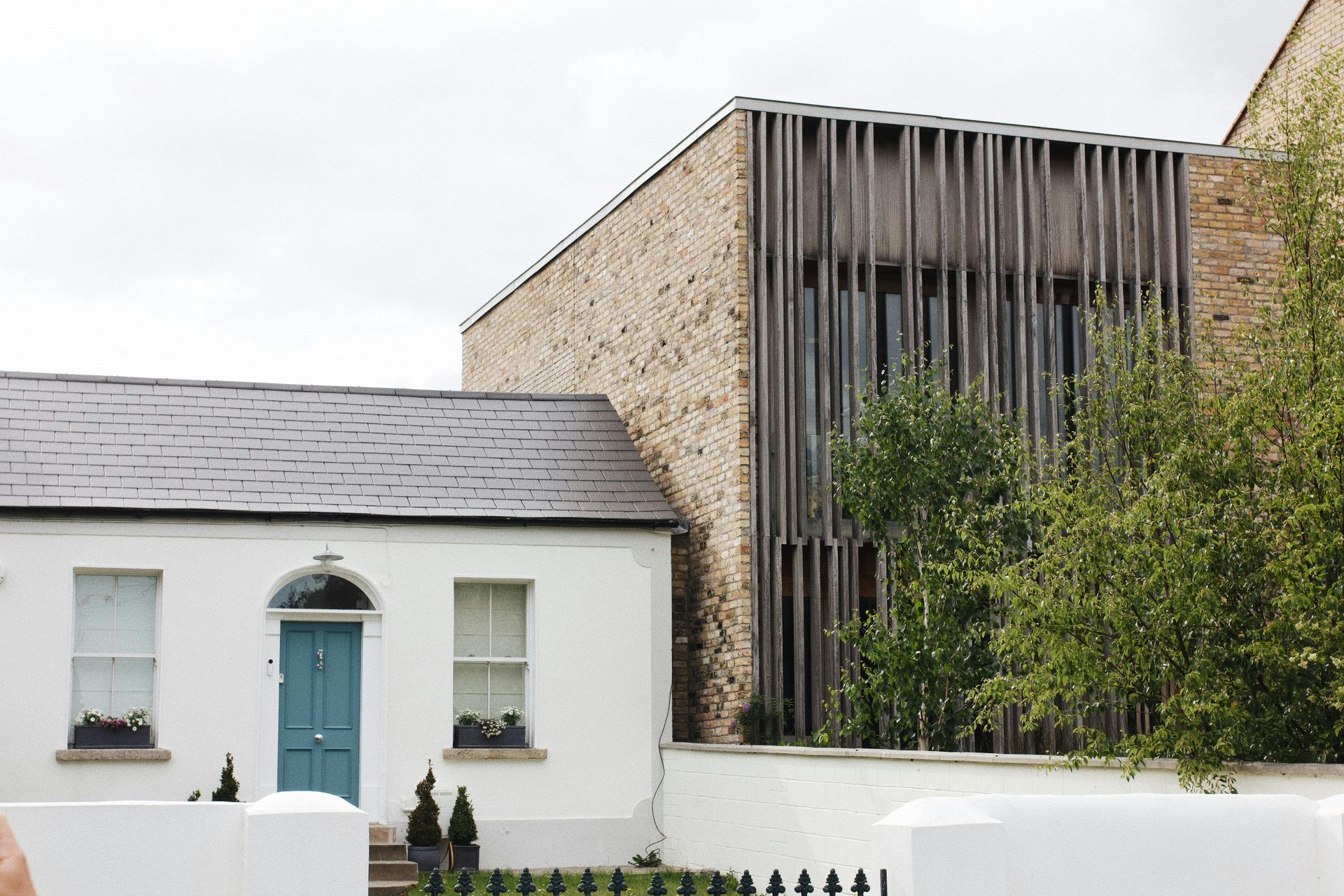 White cottage beside modern stone building with vertical slats and greenery