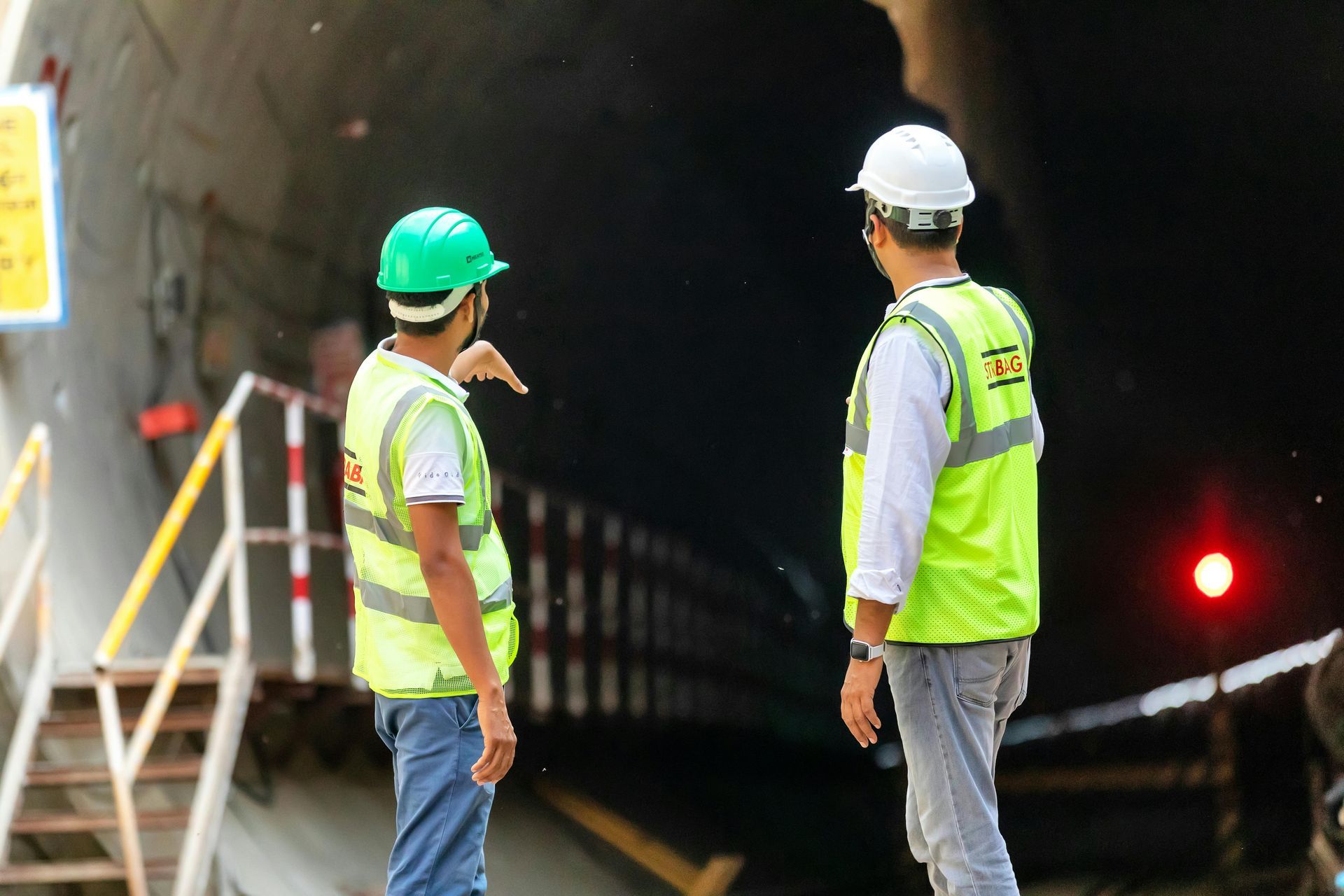 Two construction workers in safety vests and helmets stand at a tunnel entrance, facing a dark interior.