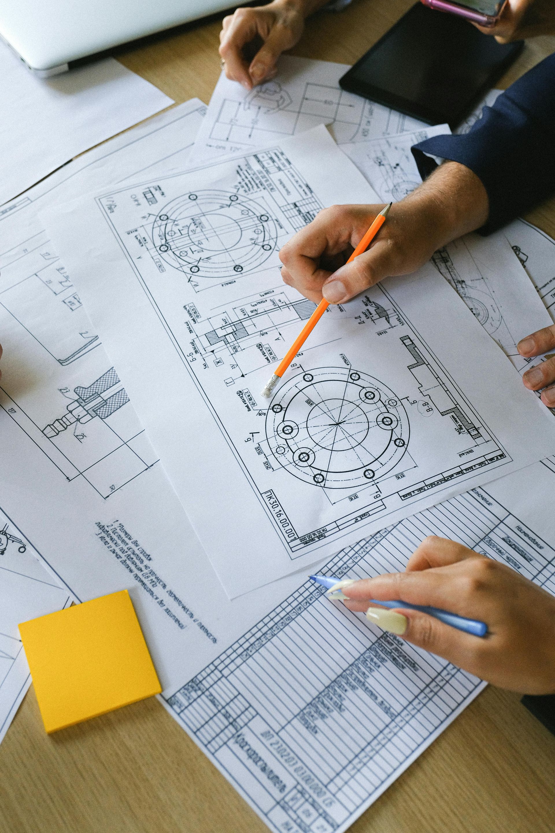 Hands reviewing technical blueprints on a desk, with pencils, papers, and a yellow sticky note.