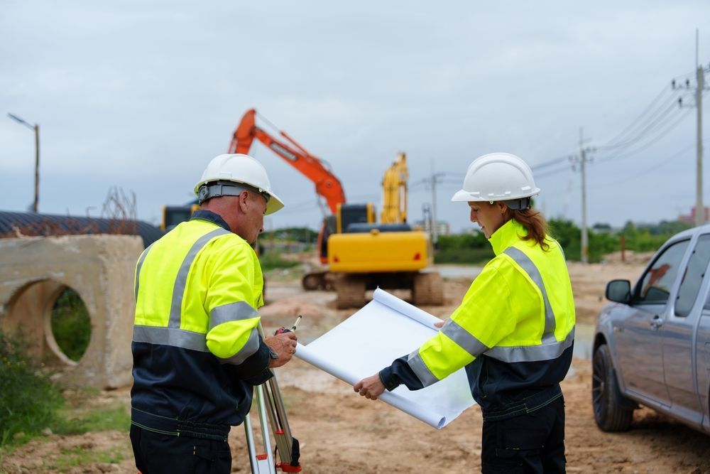 Two construction workers in safety gear reviewing blueprints at a roadside work site with excavators.