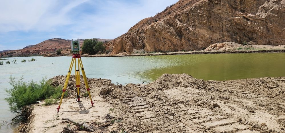 Survey tripod on rocky riverbank beside green water and brown hills under a clear sky