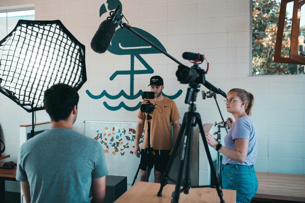 Filming a video with a camera on a tripod. Two people work behind the camera, one holding a notepad. There's a softbox, and a flamingo mural.