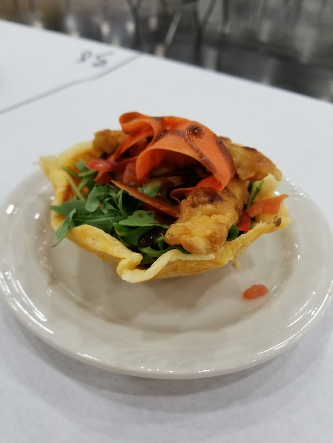 Fried food and arugula in a tortilla bowl, topped with carrot ribbons, on a white plate.