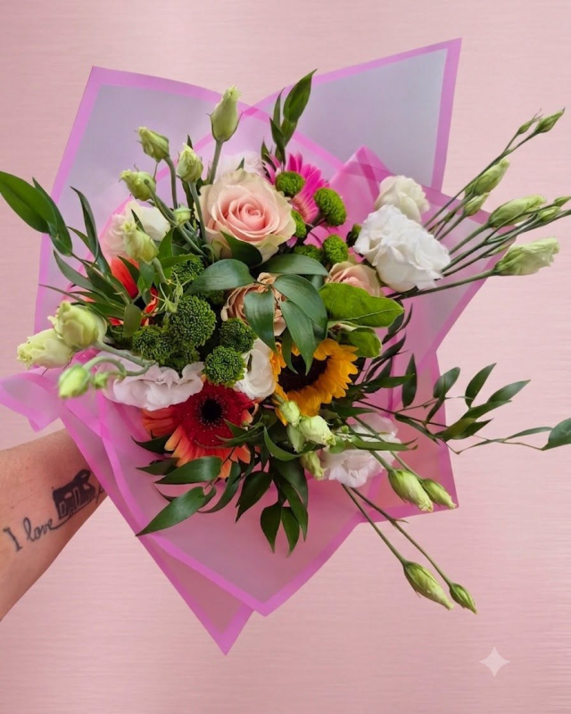 Bouquet avvolto in carta rosa con rose, girasoli, gigli e fogliame, su uno sfondo rosa