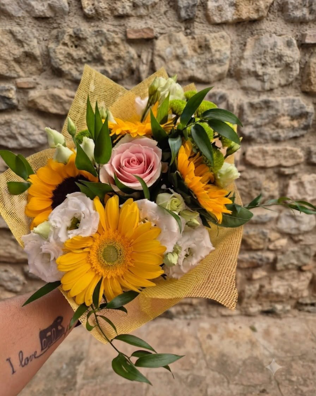 Bouquet di fiori gialli e rosa avvolto in carta marrone, davanti a un muro di pietra