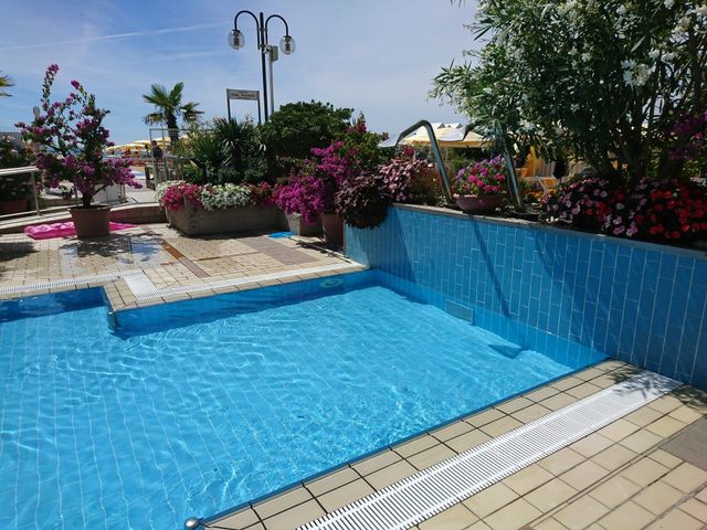Piscina blu con bordo piastrellato, fiori rosa e lampioni in un giardino soleggiato