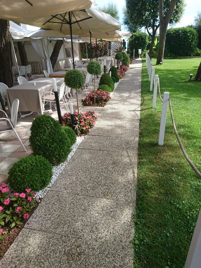Passerella in giardino accanto a un patio coperto da un tendone bianco, con tavoli, sedie, ombrelloni e piante di arte topiaria potate.