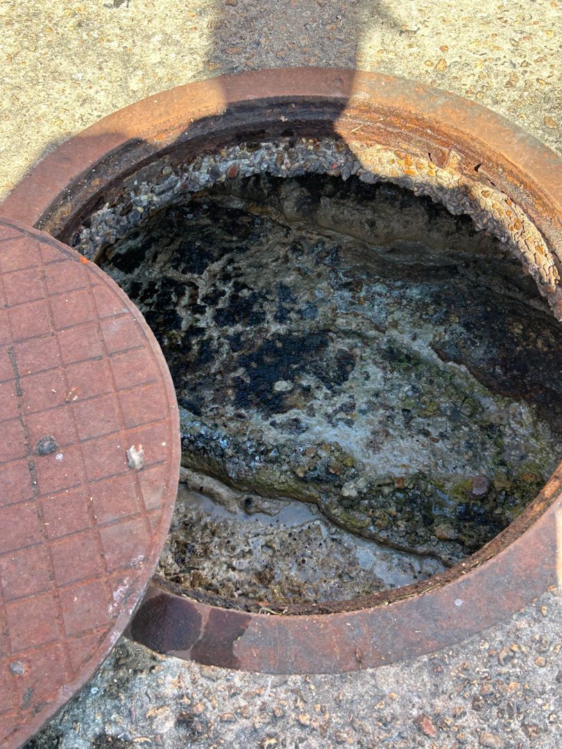 Open manhole showing a mass of congealed waste inside, a partially open rusty lid beside it.