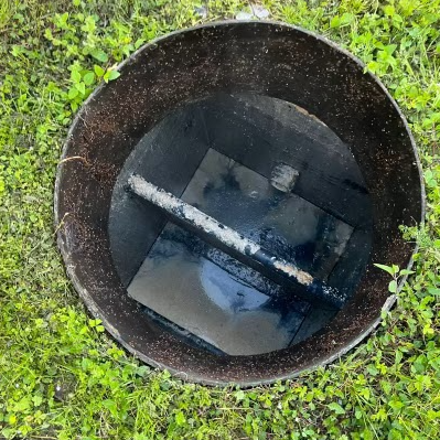 Open black sewage access hole in green grass, showing dark interior with central platform and metal bar.