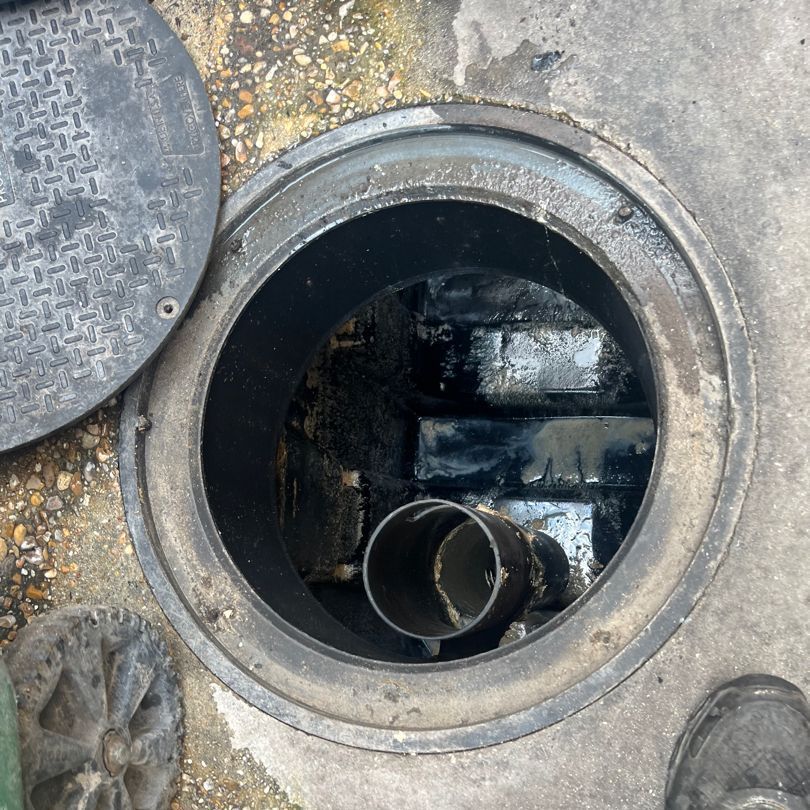 Open manhole with pipes and debris inside, on a concrete surface.