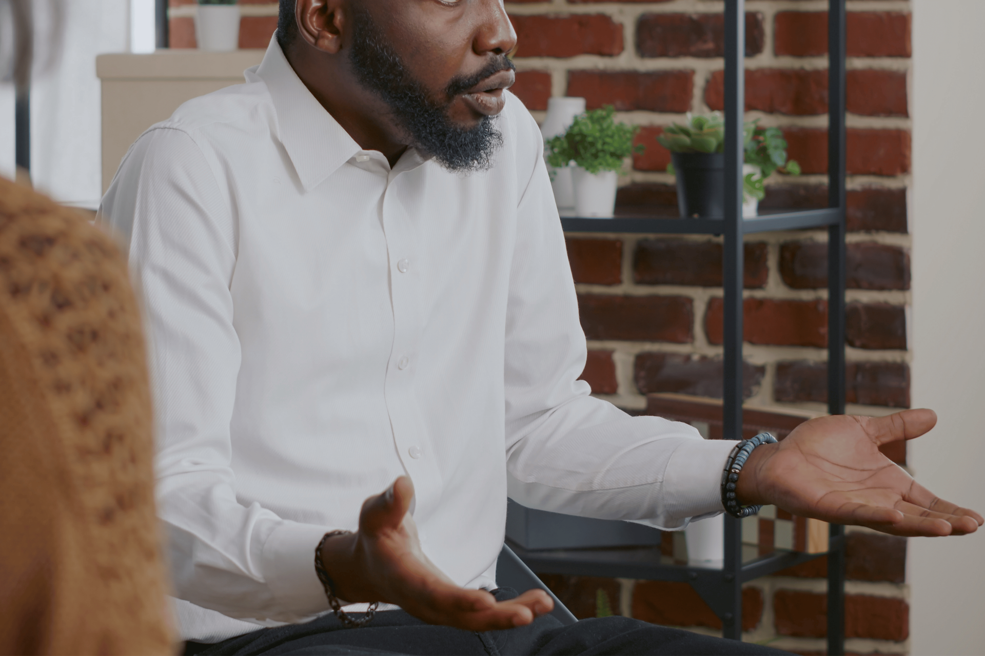 Man in a white shirt speaking with open hands in a brick-walled room with shelves and plants.
