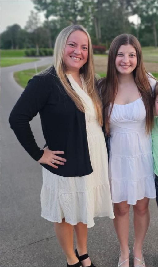 A woman in a black cardigan is standing next to two girls in white dresses.