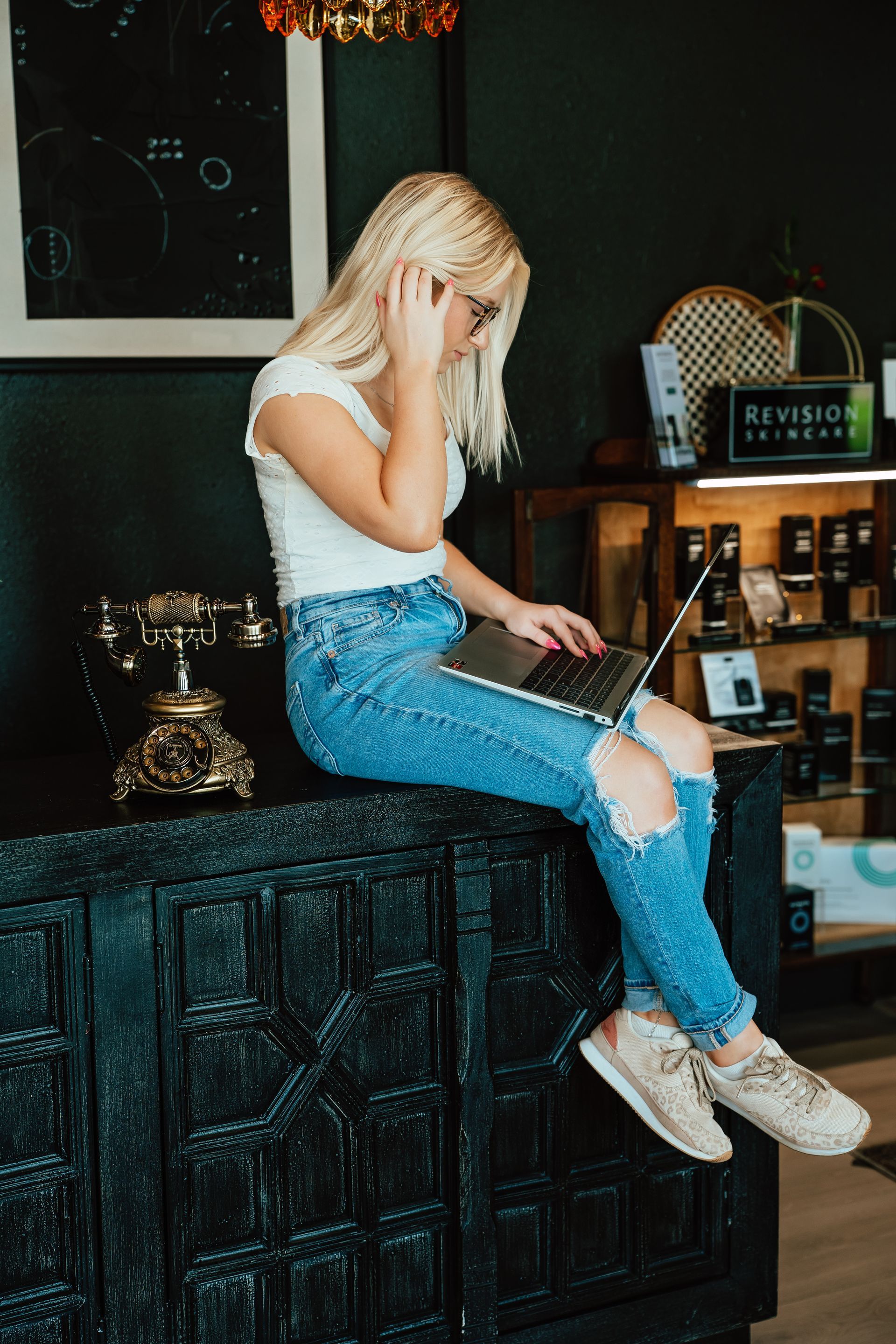 Blonde woman in ripped jeans sits on dark counter, using laptop, glasses, holding head, in shop.