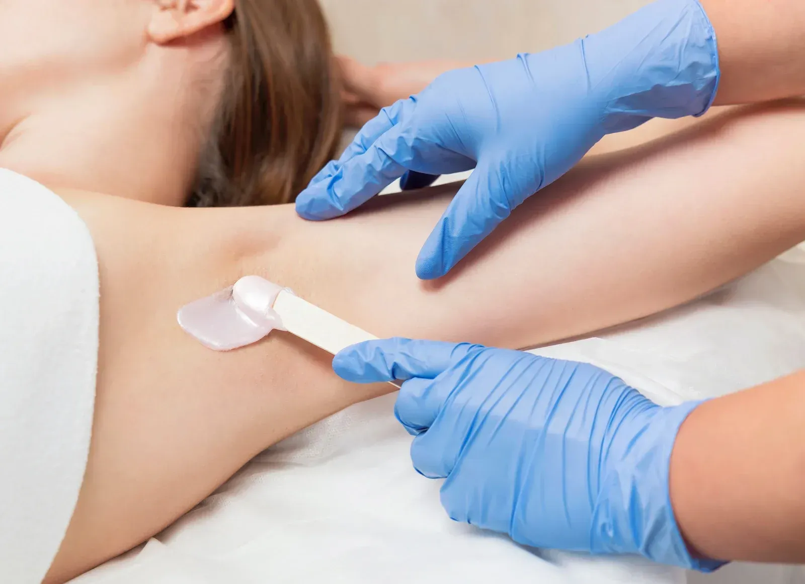 A woman is getting her armpit waxed by a woman wearing blue gloves.