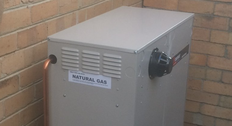 A natural gas furnace mounted outside against a brick wall. It has a vent and gas pipe.
