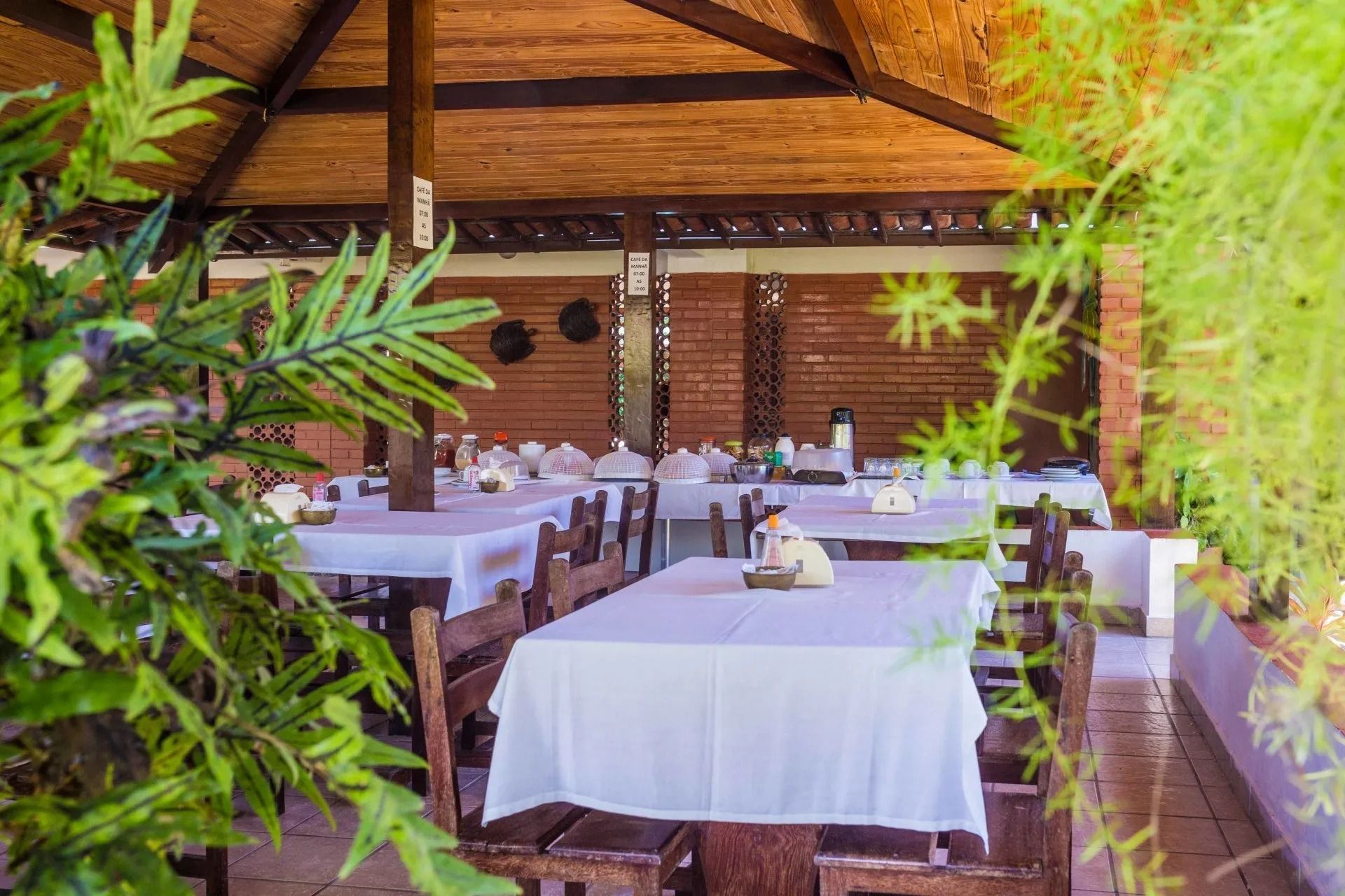Restaurante ao ar livre, com mesas cobertas por toalhas brancas sob um telhado de palha, rodeado por plantas verdes