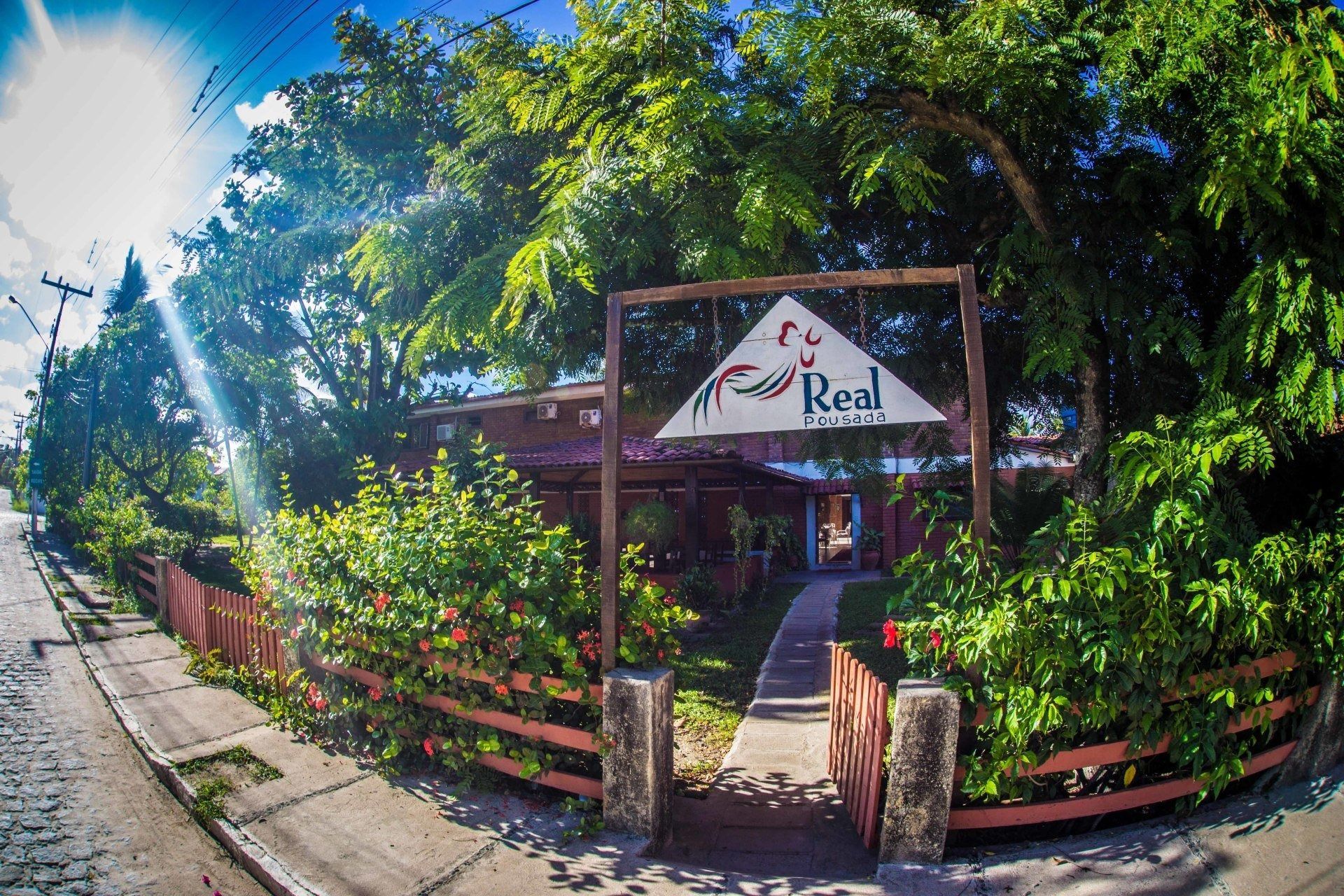 Entrada ensolarada de um café tropical, com cerca vermelha, vegetação exuberante e uma placa “Real” acima da porta