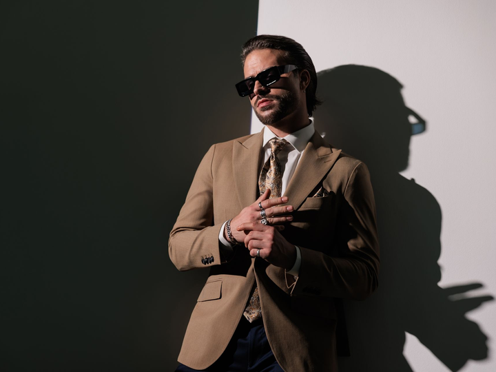 Man in tan suit and sunglasses posing against a gray wall with dramatic shadow