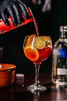A gloved hand pours a red liquid from a can into a stemmed glass filled with ice, orange slices, and a lime wheel.