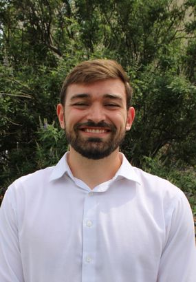 Smiling man in a white shirt standing outdoors with green trees in the background