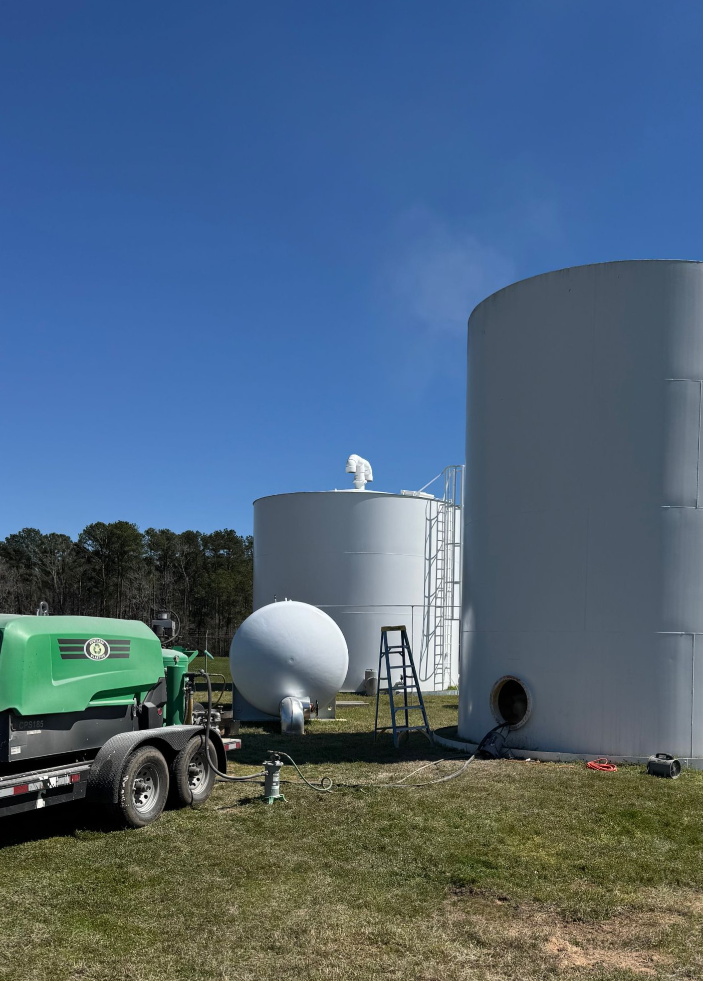 Industrial tanks and a green truck on a grassy site under a clear blue sky