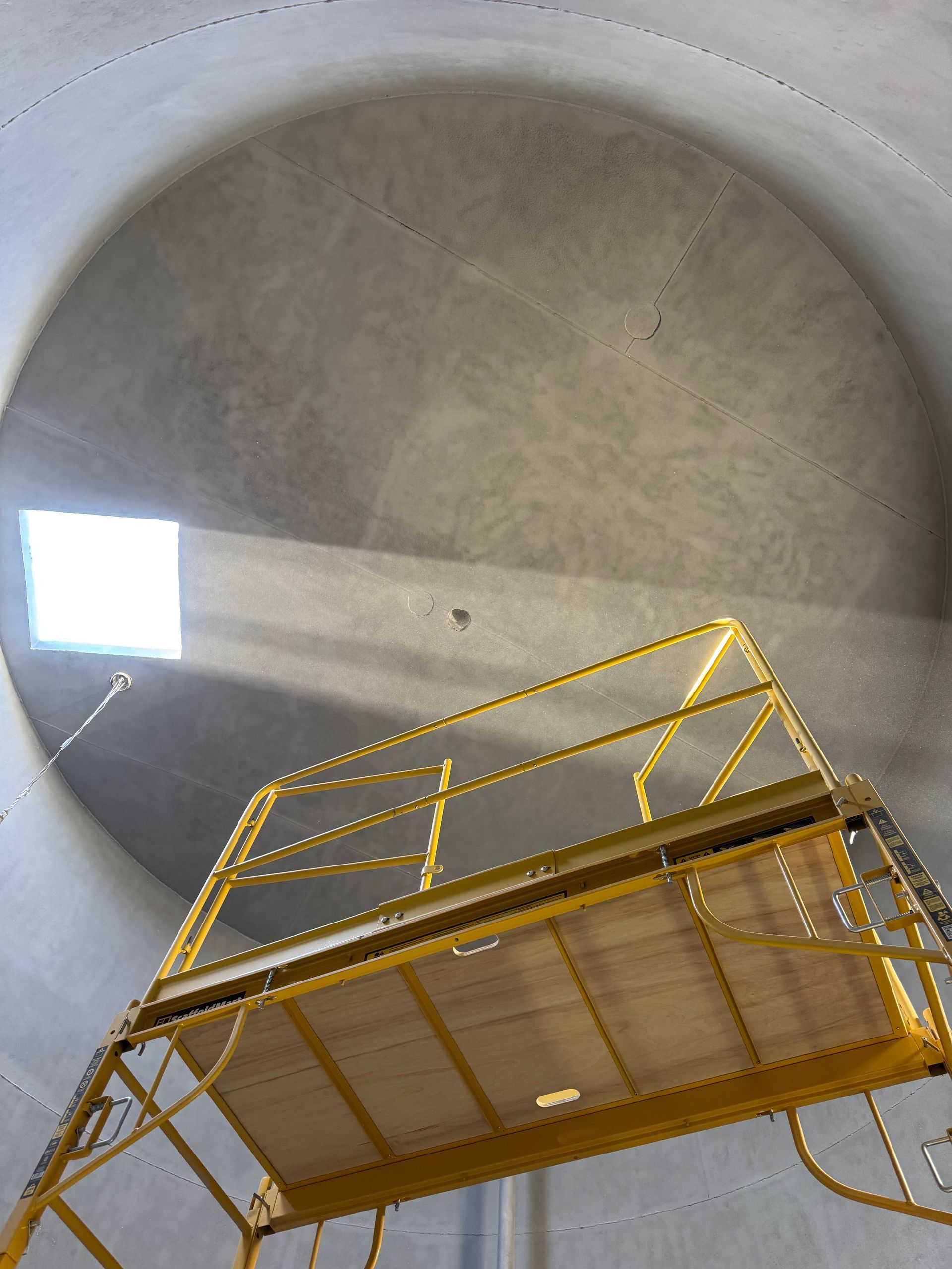 Yellow metal scaffolding inside a large concrete cylinder, viewed upward toward a bright opening.