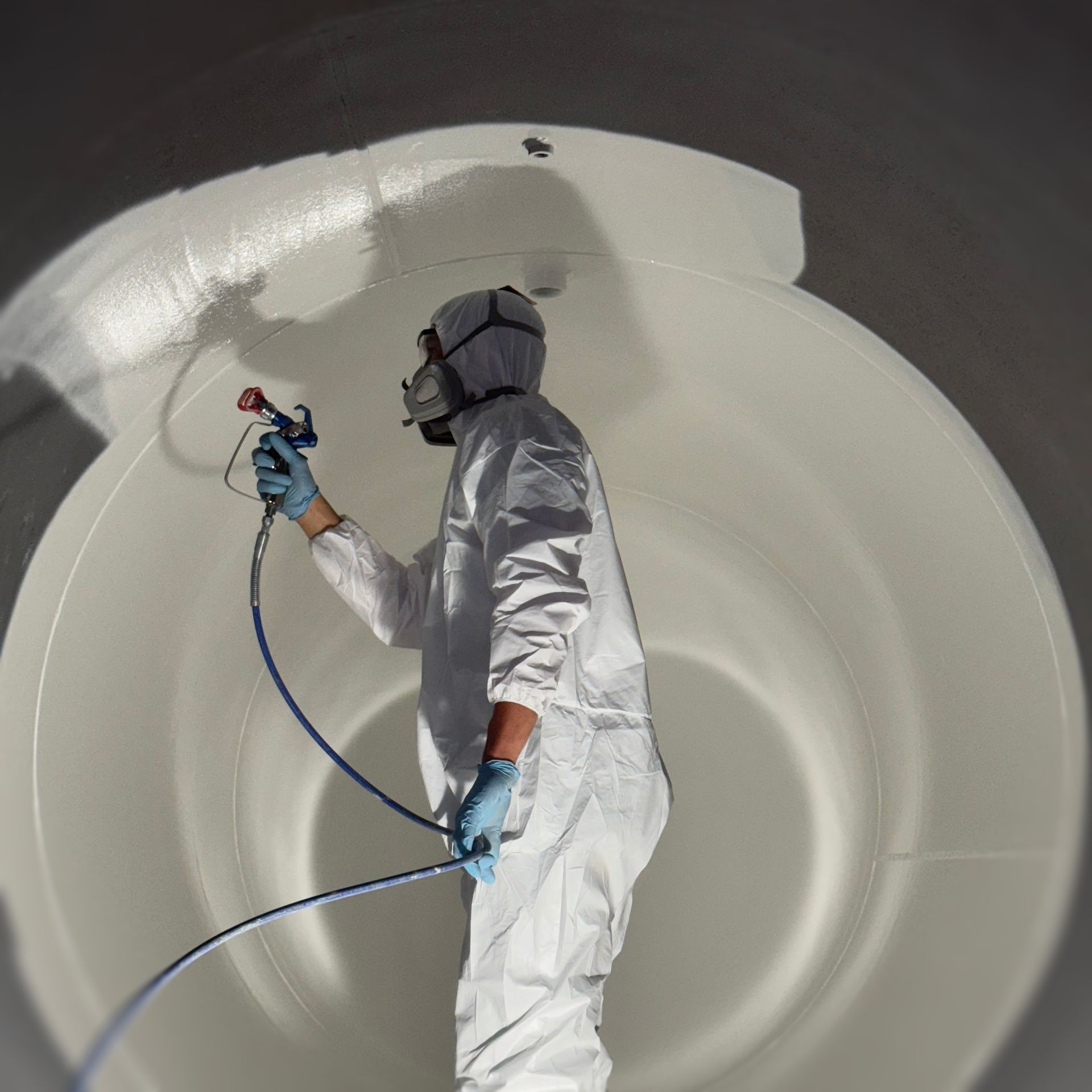Worker spraying the inside of a large white cylindrical tank with a hose and protective suit
