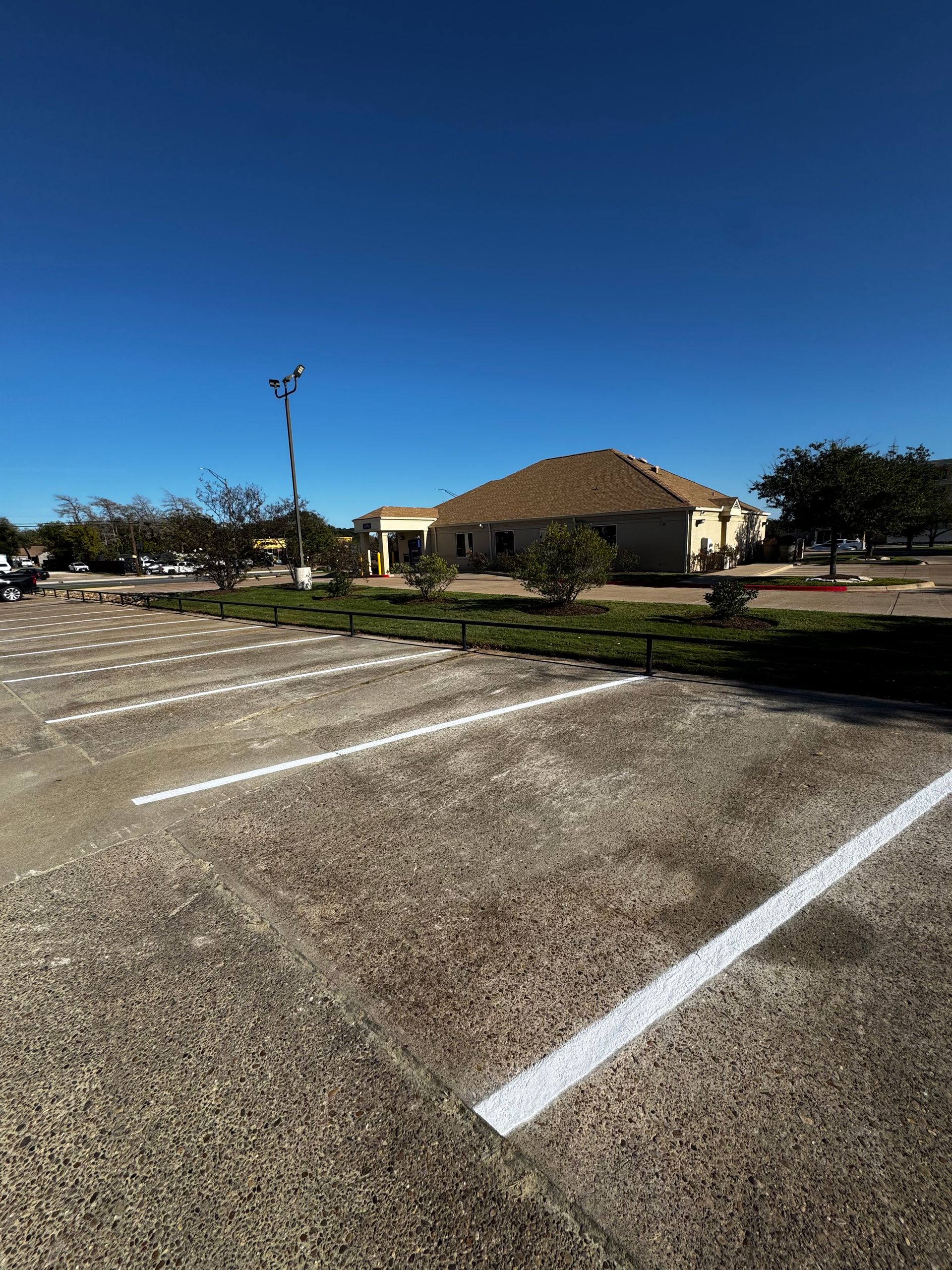 Empty parking lot with white lines, a tan building, and trees under a clear blue sky.