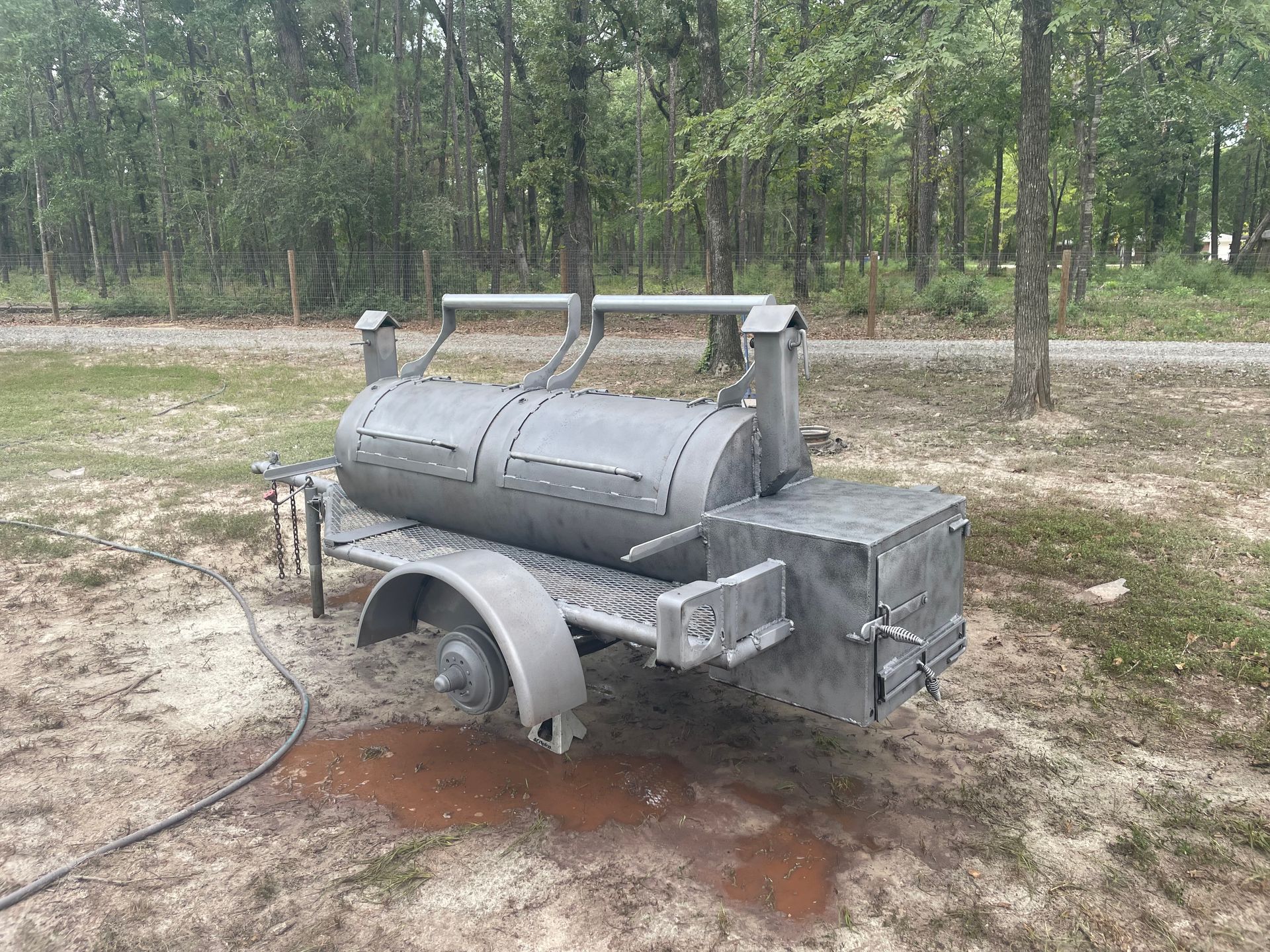 Large gray metal outdoor drum machine on a dirt clearing in a wooded area, with wheels and handlebar-like frame.