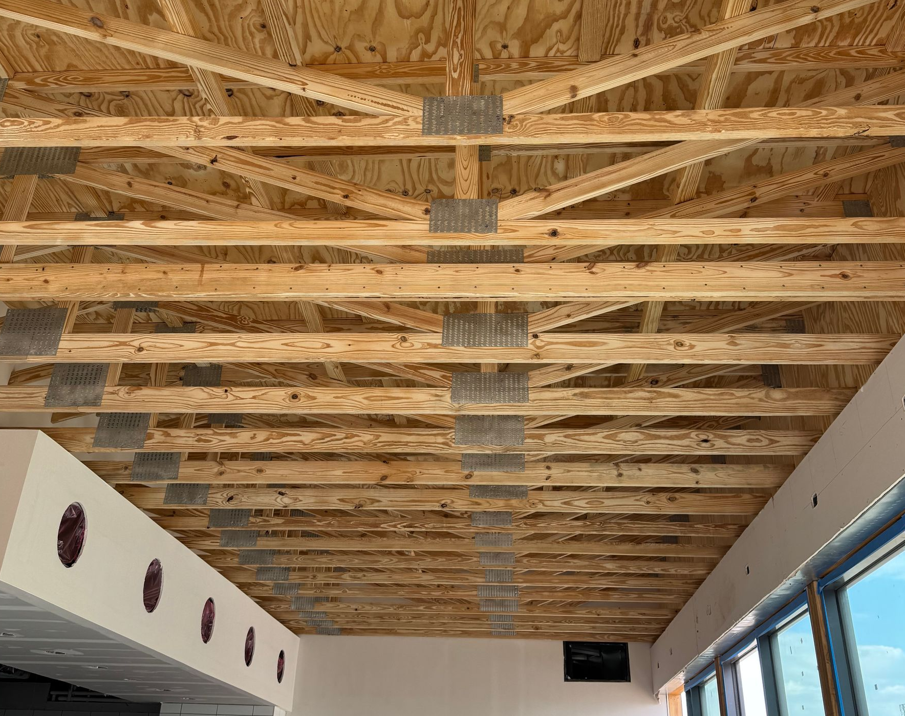 Exposed wooden ceiling trusses in an unfinished room with white walls and windows on the right.