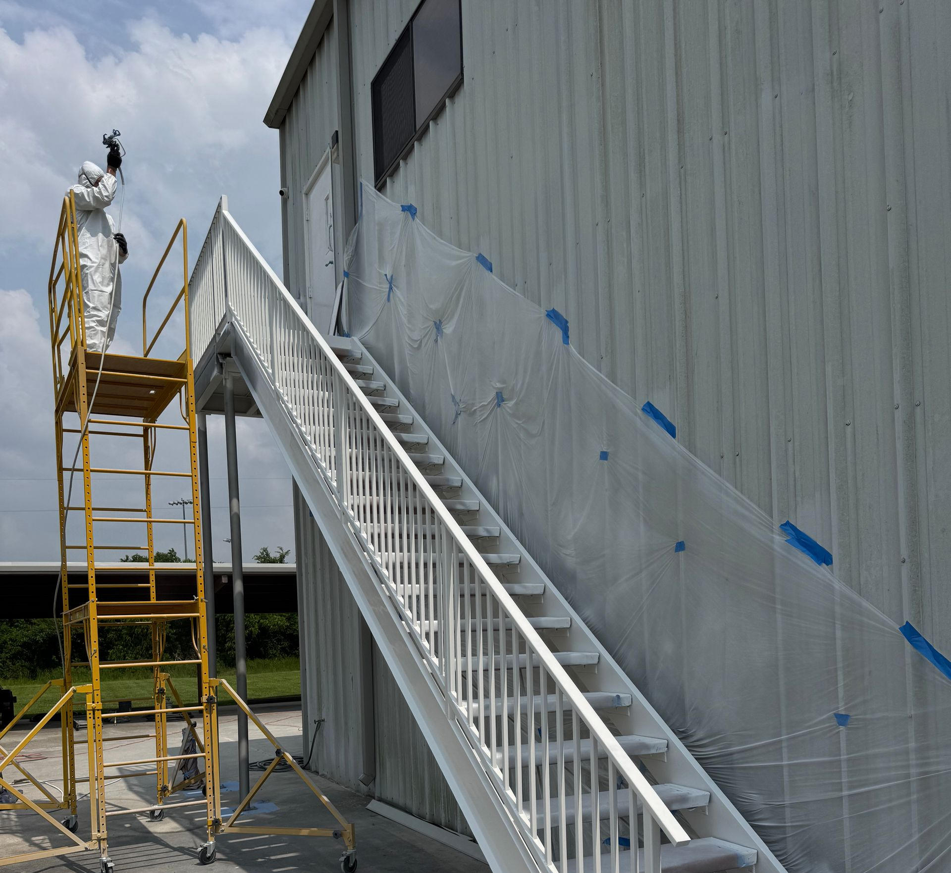 Worker on yellow scaffolding painting a gray building beside a white stairway.