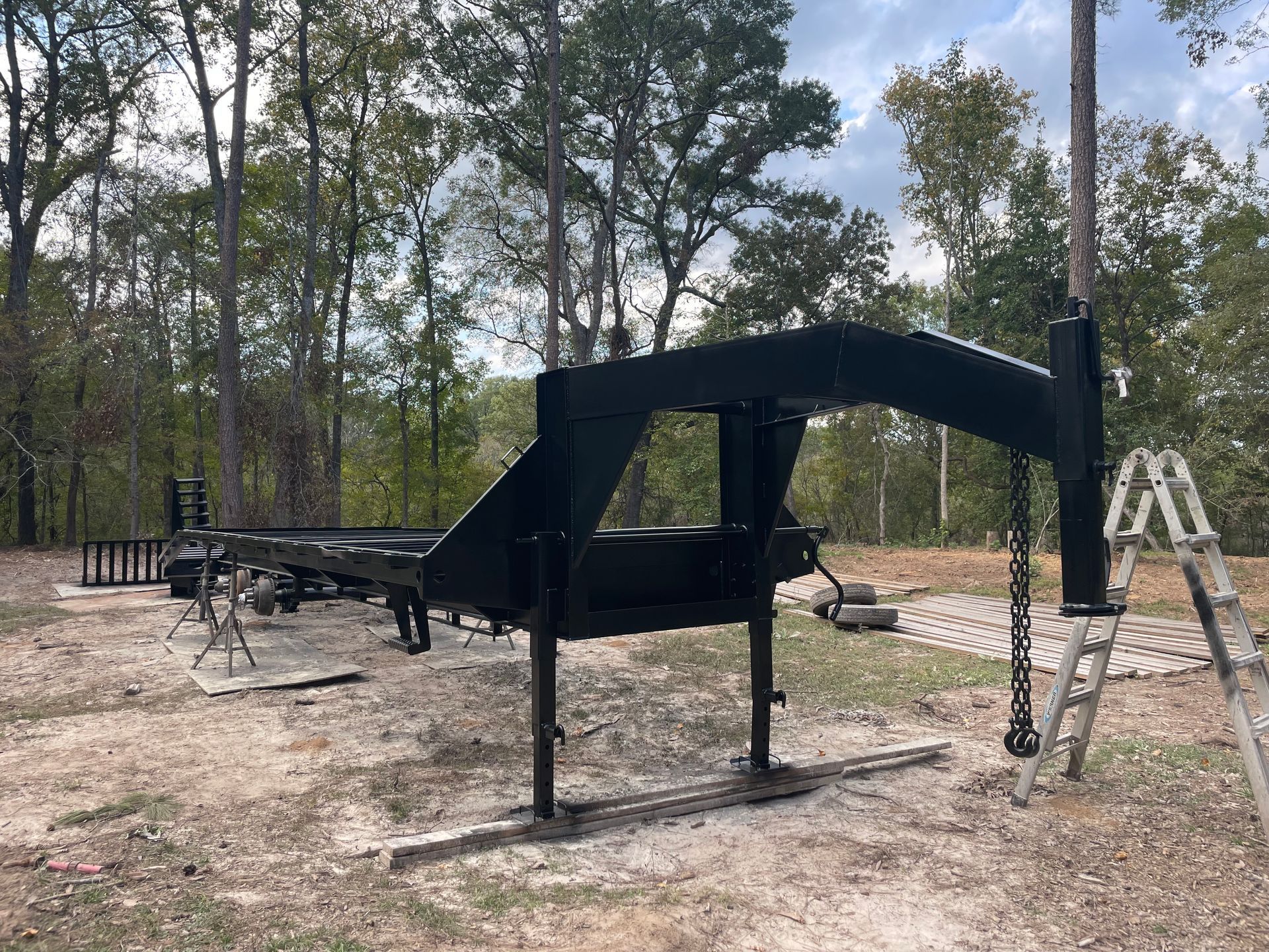 Black utility trailer frame on a dirt lot, with a ladder nearby and trees in the background