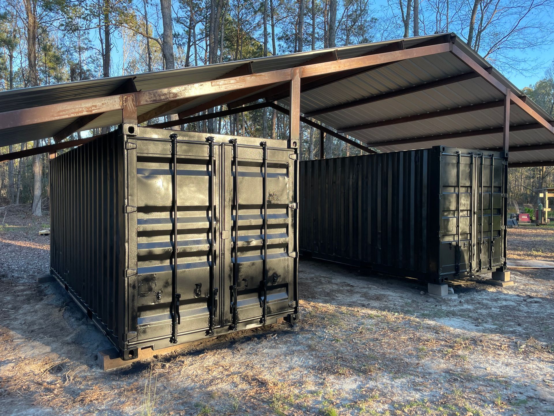 Two black shipping containers under a metal roof in a wooded outdoor lot