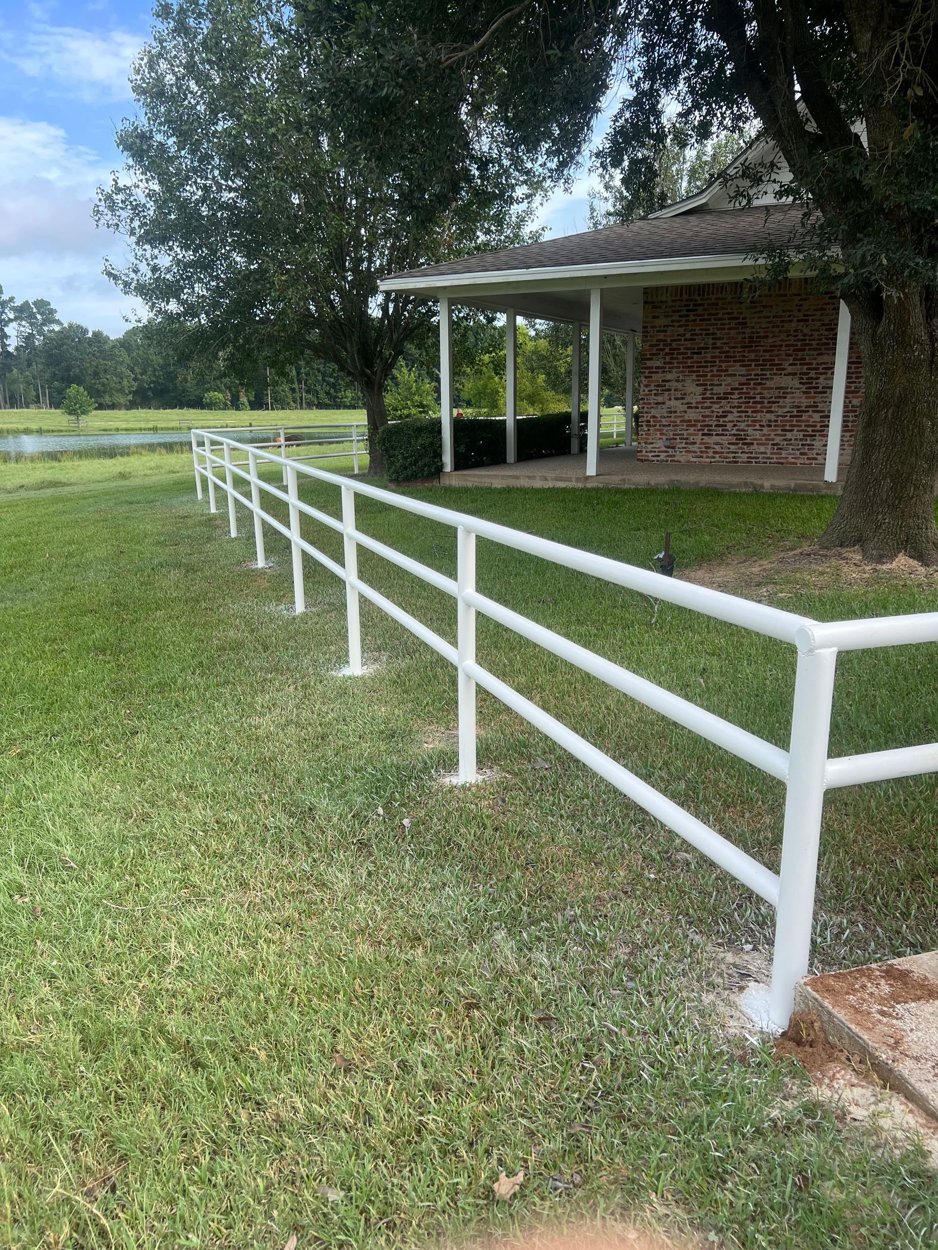 White rail fence beside a small brick shed on a grassy lawn with trees in the background