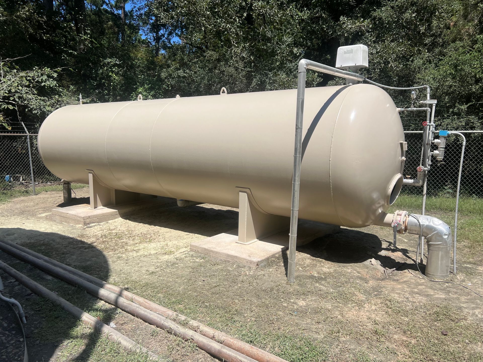 Large tan cylindrical tank on concrete supports outdoors, with pipes and a valve assembly beside it.