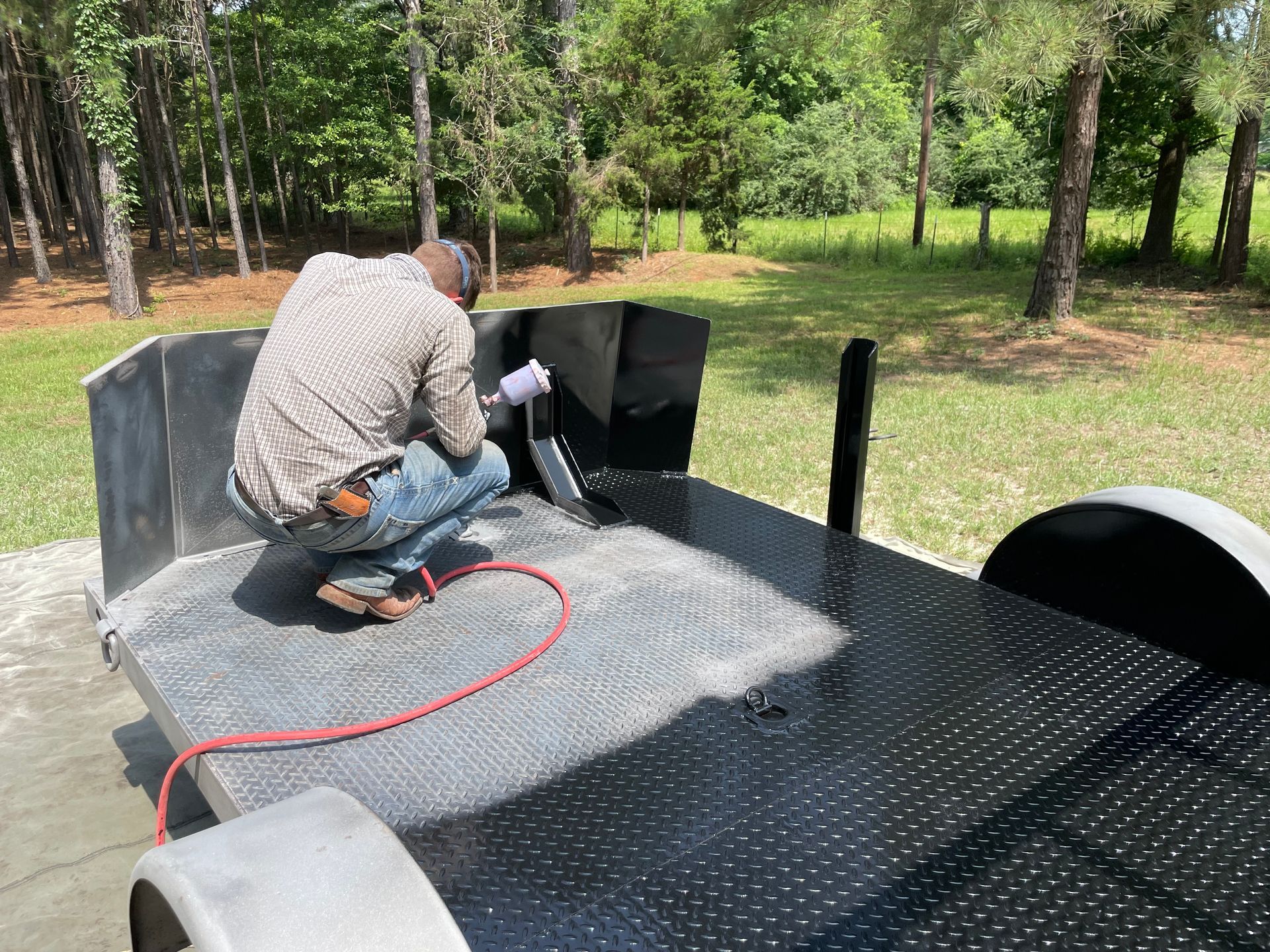 Person kneeling on a truck bed liner, spraying or coating the surface outdoors in a wooded area.