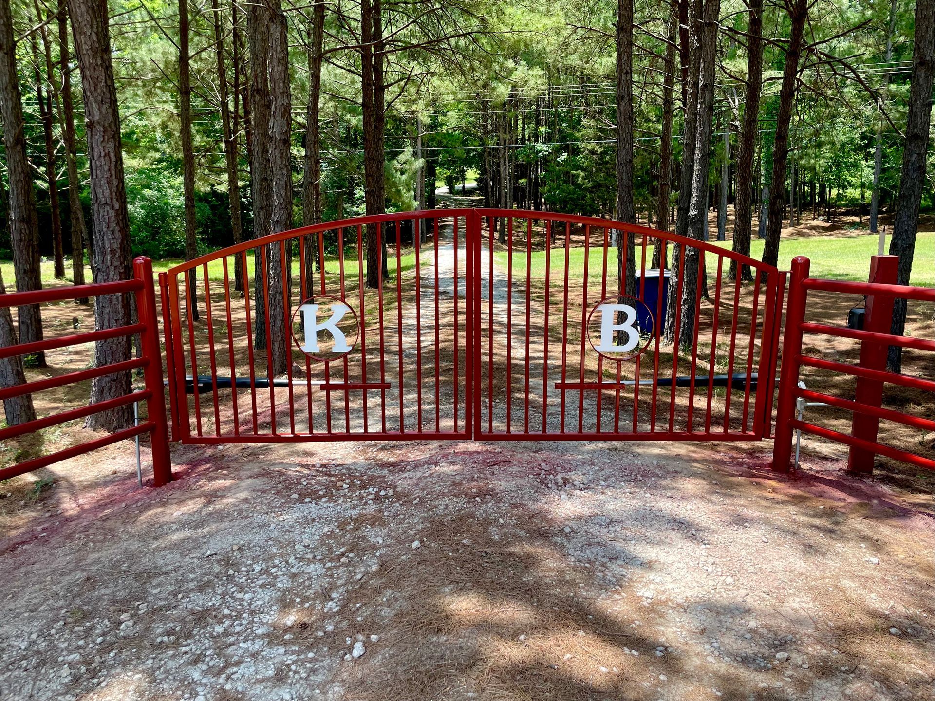 Red metal gate with white letters B and B at a wooded entrance, framed by trees and gravel path