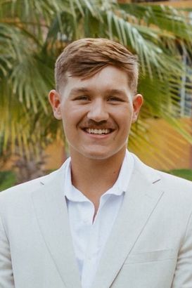 Smiling person in a white blazer standing outdoors with palm trees in the background