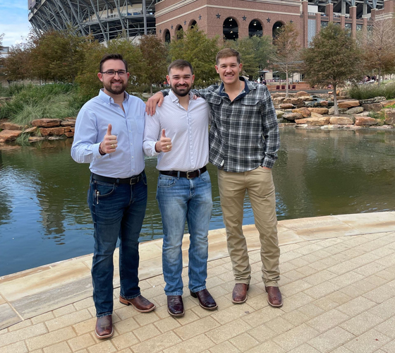 Three people standing by a pond, smiling and giving thumbs up in front of a stadium.