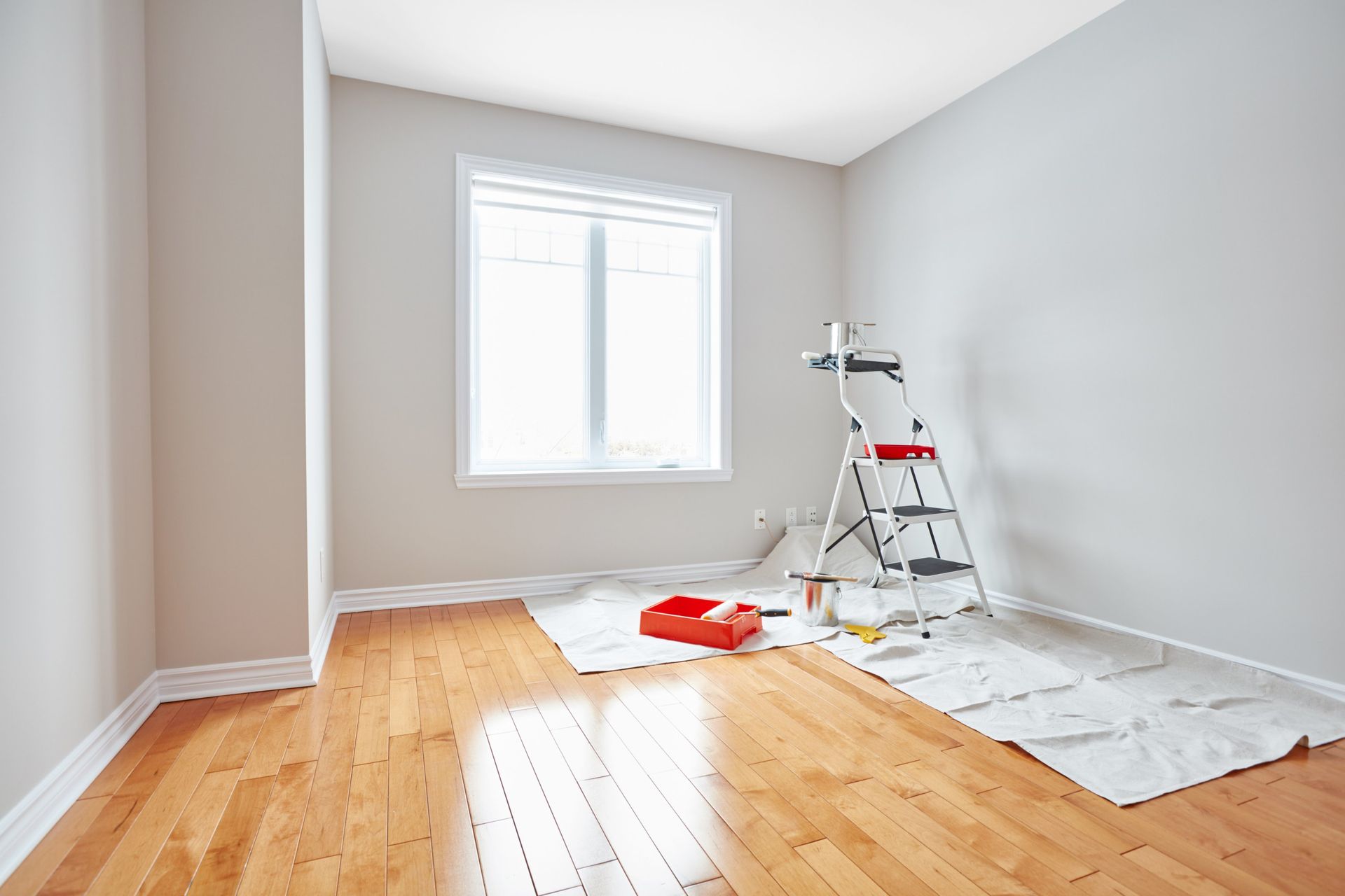 Empty room being painted, paint supplies on floor and ladder. Light-colored walls and hardwood floor.