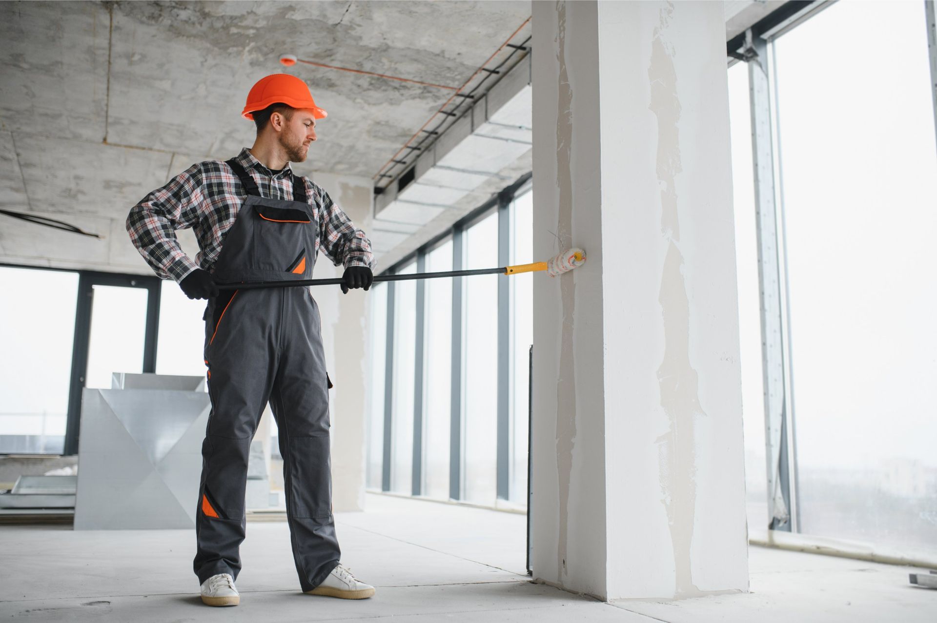 Construction worker in hard hat, painting a column with a roller in a building under construction.