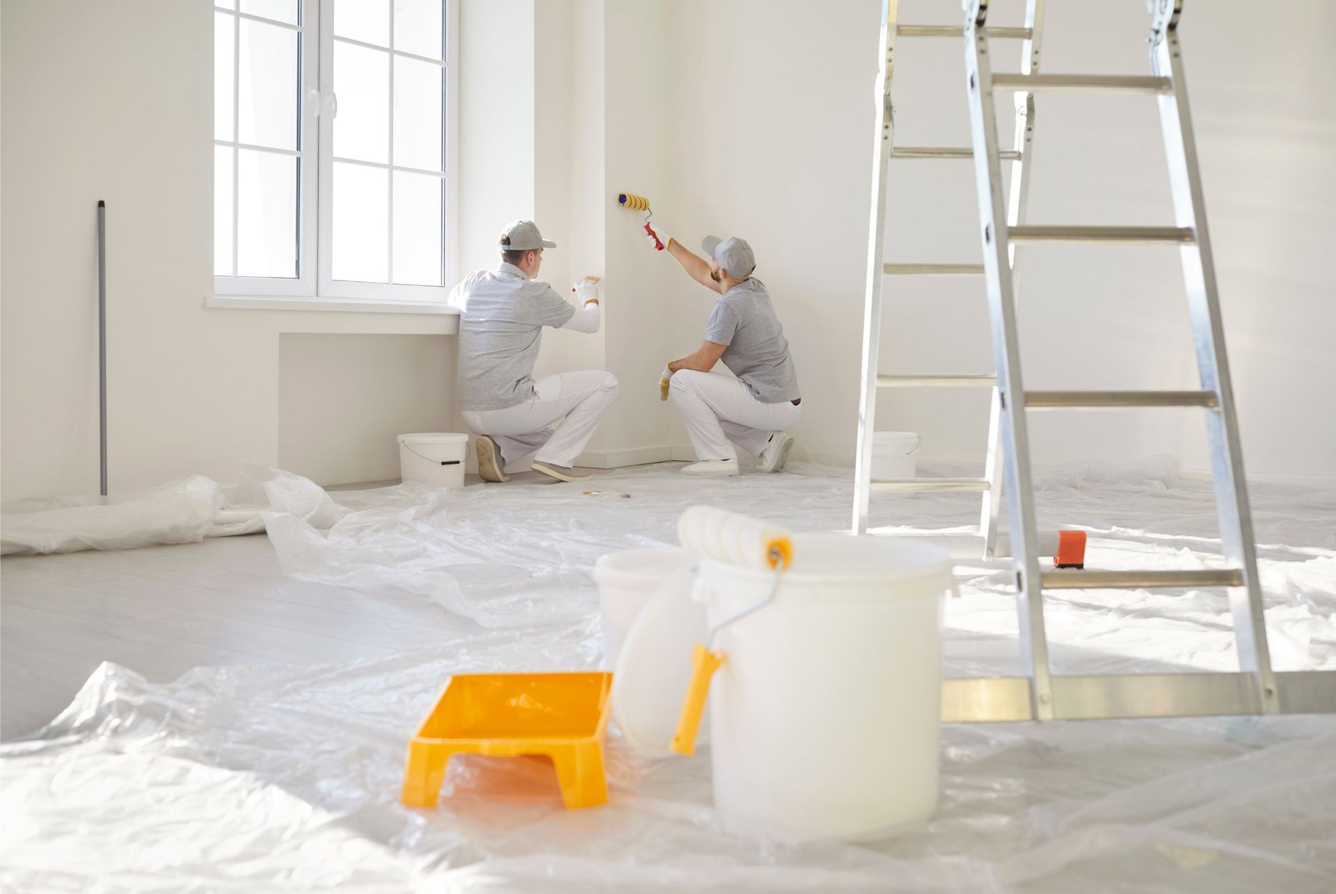 Two people painting a white wall in a room. Paint rollers, ladder, paint buckets, and drop cloths are present.
