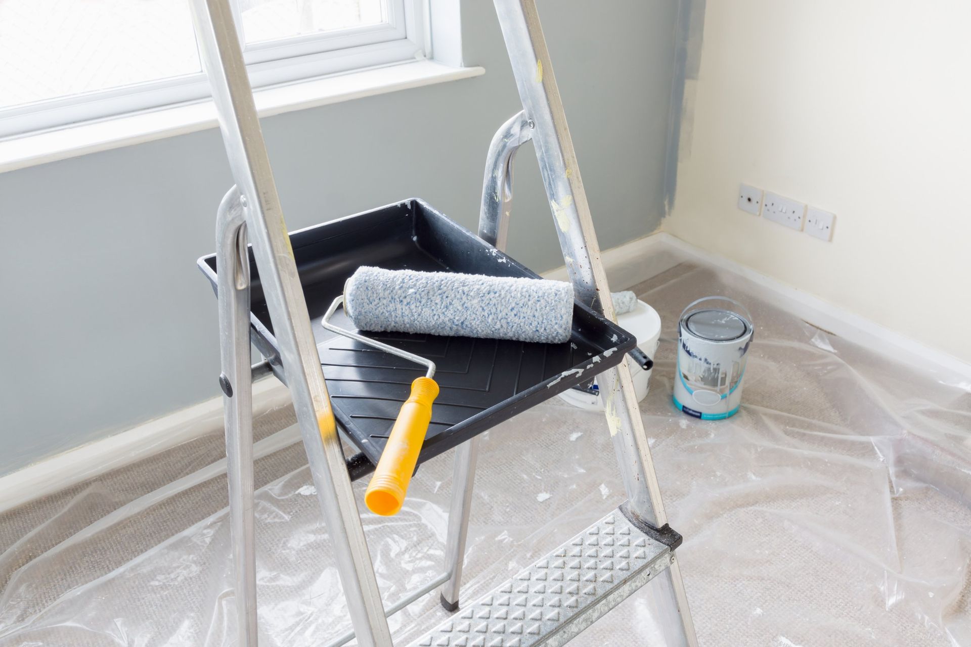 Ladder with paint roller and tray, near a freshly painted gray wall.