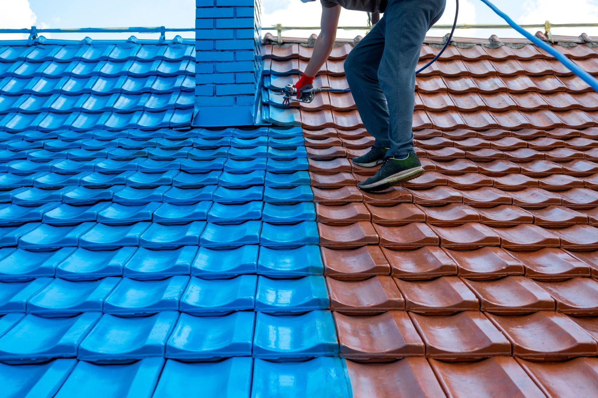 A person painting roof tiles blue from a rooftop. Half of the roof is blue, the other half is brown.