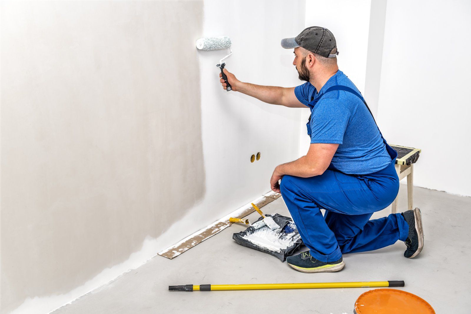 Painter in blue overalls kneels, rolling white paint on a wall. Tools and paint tray on the floor.